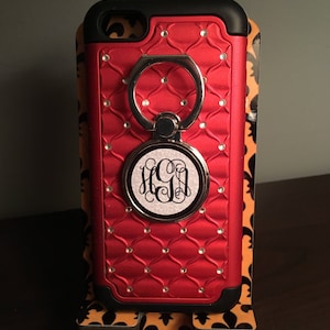 May include: Red phone case with a quilted pattern, a silver ring holder, and a monogram. The case has a black border and is embellished with small, clear rhinestones. The phone case is displayed on a decorative stand.