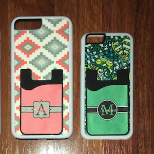 May include: Two white phone cases with a pocket for a credit card. The case on the left has a geometric pattern in shades of gray, white, and red. The case on the right has a green, blue, and yellow floral pattern. Both cases have a black pocket with a personalized monogram.