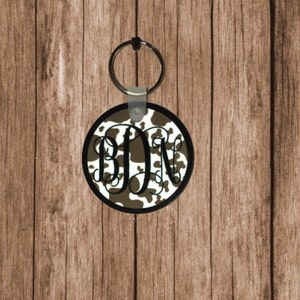 May include: A round, brown and white cowhide patterned keychain with a black metal ring and a black and white monogram "BDN" in the center.