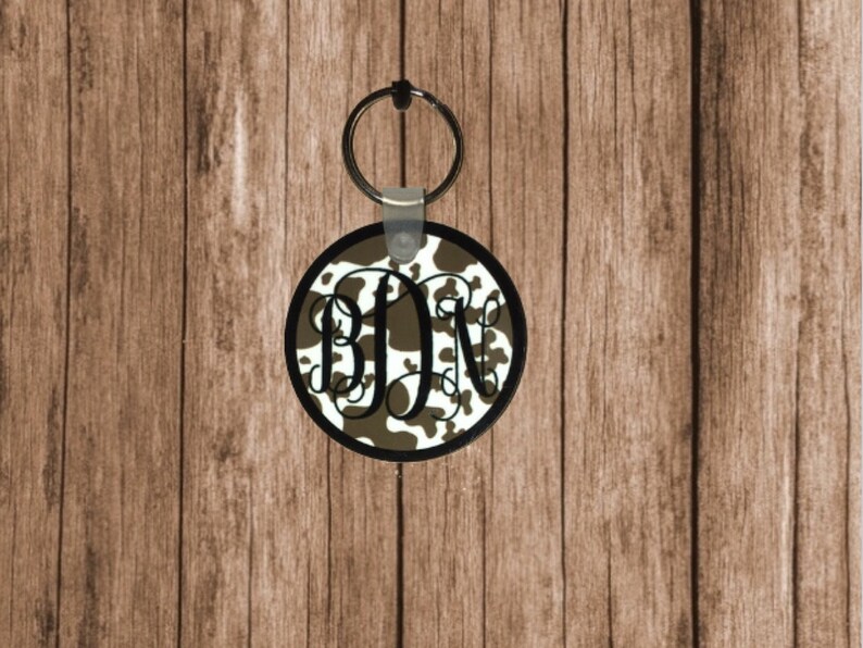 May include: A round, brown and white cowhide patterned keychain with a black metal ring and a black and white monogram "BDN" in the center.