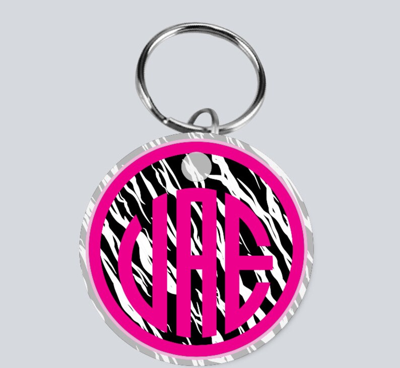May include: A round keychain with a black and white zebra print background. The keychain has a pink circle with the letters "VAE" in white inside the circle.