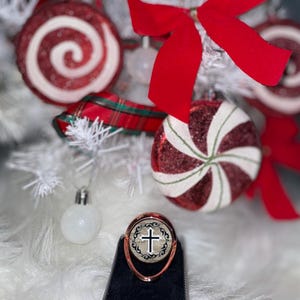 May include: A rose gold phone ring holder with a black and white design featuring a cross. The ring holder is on a black surface with a white fuzzy background.