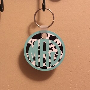 May include: A round keychain with a black and white cow print design. The keychain has a turquoise monogram in the center. The keychain is attached to a silver key ring.