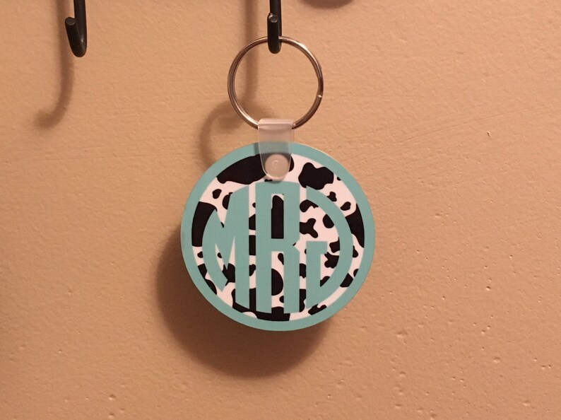 May include: A round keychain with a black and white cow print design. The keychain has a turquoise monogram in the center. The keychain is attached to a silver key ring.