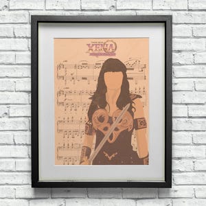 Op de afbeelding: Een ingelijste print van een vrouw met lang donker haar die een zwaard vasthoudt. De print is op een blad muziek met de titel "Theme from Xena: Warrior Princess".
