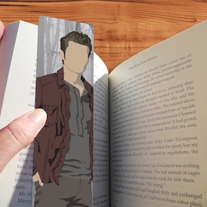 May include: A brown and gray illustrated bookmark featuring a man with a dark brown jacket and gray shirt. The bookmark is set against a background of trees. The text "Over By Cate" is visible on the right side of the bookmark.
