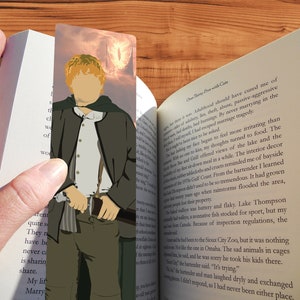 May include: A bookmark featuring a cartoon illustration of a red-haired character wearing a brown jacket and holding a sword. The background is a fiery orange and red with a large, glowing eye in the distance.
