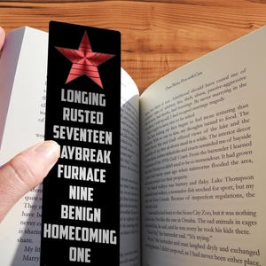 Peut inclure: Un marque-page en métal noir avec une étoile rouge et les mots "Longing", "Rusted", "Seventeen", "Daybreak", "Furnace", "Nine", "Benign", "Homecoming", et "One" en texte blanc.