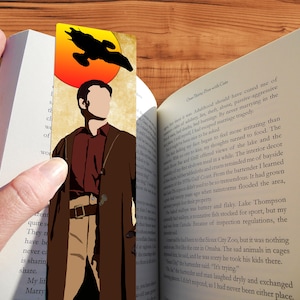Könnte beinhalten: Ein braunes und beiges Lesezeichen mit einer Silhouette eines Raumschiffs, das über einer untergehenden Sonne fliegt. Das Lesezeichen zeigt einen Charakter aus der Serie "Firefly", der vor der Sonne steht. Das Lesezeichen befindet sich in einer Hand, die ein Buch hält.