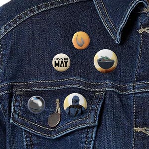 Könnte beinhalten: Eine Jeansjacke mit fünf verschiedenen Anstecknadeln. Die Nadeln sind rund und zeigen verschiedene Figuren aus dem Star Wars-Universum. Die Nadeln zeigen einen Mandalorianer, einen Baby Yoda, einen Charakter aus der Mandalorianer-Serie und einen Charakter aus der Star Wars-Serie.