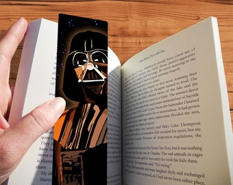 May include: A black and orange bookmark featuring a stylized illustration of Darth Vader from Star Wars. The bookmark has a dark background with a subtle sparkle effect.