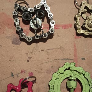 May include: A collection of keychains crafted from recycled bicycle chains and metal hardware components. The items feature various shapes including hearts and circular designs, finished in colors such as metallic silver, bright pink, lime green, and gold.