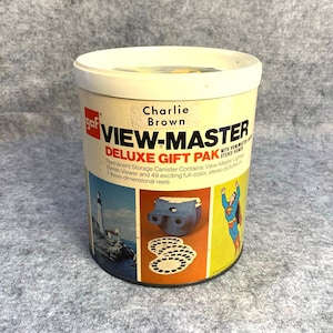 May include: A vintage View-Master Deluxe Gift Pak canister with a white lid and a cylindrical body. The canister features the text "Charlie Brown" and illustrations of a lighthouse, a stereo viewer, and Superman.