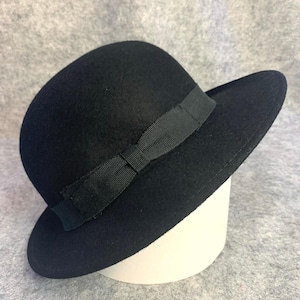 May include: A black cloche hat with a rounded crown and a small brim. A black ribbon is wrapped around the base of the crown and tied in a bow. The hat is displayed on a white cylindrical form.