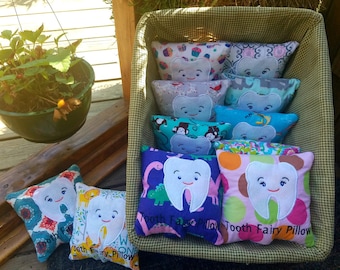 Op de afbeelding: Een mand vol kleurrijke tandenfee kussens. Elk kussen heeft een witte tand met een glimlachend gezicht en verschillende patronen. De kussens zijn gelabeld met "Tooth Fairy Pillow".