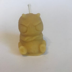 May include: A small, yellow, beeswax candle shaped like a cartoon monster with a grumpy expression. The candle has a white wick.