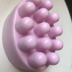 May include: A lavender-colored bath soap with a textured surface. The soap has multiple rounded bumps on the top.