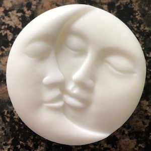 May include: White ceramic sculpture of two crescent moons facing each other, with closed eyes and noses touching. The sculpture is a modern interpretation of the moon.
