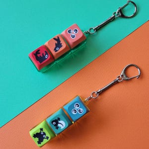 May include: Two keychains featuring three square, colourful keycap-style blocks each. The top keychain has red, orange, and pink blocks with black fish and swirl graphics. The bottom keychain has lime, teal, and blue blocks with black frog and swirl graphics.