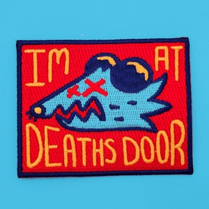 Puede incluir: Parche bordado rojo con un monstruo de dibujos animados azul y azul claro con una X sobre su ojo. El texto "I'M AT DEATHS DOOR" está en bordado amarillo.