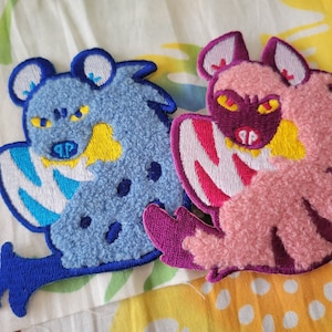 Puede incluir: Dos parches bordados de felpa, uno azul con detalles blancos y amarillos, el otro rosa con detalles blancos, amarillos y morados. Ambos parches presentan un animal de dibujos animados con una expresión feroz.