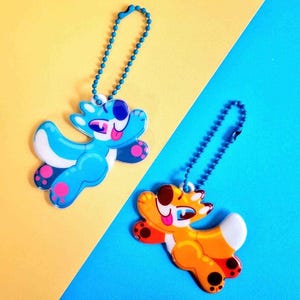Peut inclure: Deux porte-cl&eacute;s en forme d'animaux de dessin anim&eacute; color&eacute;s. Le porte-cl&eacute;s bleu pr&eacute;sente un animal bleu et blanc avec une langue rose qui sort. Le porte-cl&eacute;s orange pr&eacute;sente un animal orange et blanc avec une langue rose qui sort.