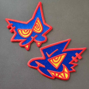 Könnte beinhalten: Zwei gestickte Aufnäher mit einem cartoonartigen Monsterdesign. Die Aufnäher haben einen blauen Hintergrund mit roten und gelben Akzenten. Das Monster hat eine sternartige Form mit einem breiten, zahnigen Grinsen und stilisierten Augen.