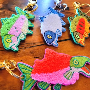 Könnte beinhalten: Gestickte Fisch-Schlüsselanhänger in verschiedenen Farben, darunter Grün, Rosa, Orange und Blau. Jeder Schlüsselanhänger hat ein detailliertes Fischdesign mit einem goldfarbenen Schlüsselring. Die Schlüsselanhänger sind auf einer Holzoberfläche angeordnet.