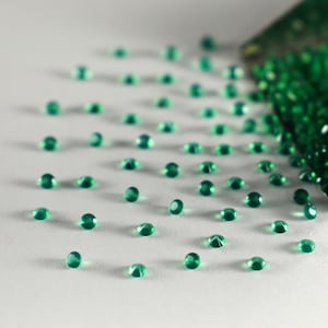 May include: Emerald green gemstones scattered on a white surface, with a small metal scoop overflowing with more gems. The gemstones are round and faceted, reflecting light. The image suggests a collection of loose gemstones.