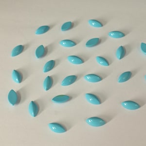 Könnte beinhalten: Eine Ansammlung kleiner, türkisfarbener, oval geformter Perlen auf einer weißen Oberfläche. Die Perlen haben eine glänzende Oberfläche und sind gleichmäßig verteilt, wodurch ein ansprechendes Muster entsteht. Die Gesamtästhetik ist sauber und minimalistisch.