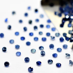 Puede incluir: Primer plano de pequeñas gemas azules redondas y facetadas dispersas. Una parte de las gemas se derrama de una cuchara blanca. Las gemas son de un color azul intenso y rico, algunas parecen más claras.