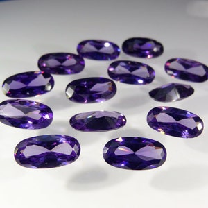 May include: A collection of twelve oval-shaped, faceted amethyst gemstones. The gemstones are a deep purple color and have a high shine.
