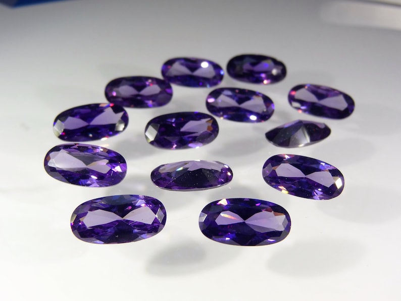 May include: A collection of twelve oval-shaped, faceted amethyst gemstones. The gemstones are a deep purple color and have a high shine.