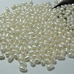Könnte beinhalten: Ein großer Haufen weißer, ovaler Süßwasserperlen. Die Perlen sind auf einer weißen Oberfläche verstreut und haben verschiedene Größen.