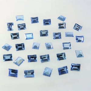 Puede incluir: Una colección de pequeñas piedras preciosas azules rectangulares y facetadas esparcidas sobre una superficie lisa. Cada piedra presenta un tono azul claro transparente con cortes geométricos precisos que reflejan la luz de forma uniforme.