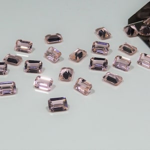 Puede incluir: Una colección de piedras preciosas sueltas de corte esmeralda con un tono rosa violáceo pálido esparcidas sobre una superficie. Una pequeña pala de metal está colocada en la parte superior derecha, conteniendo piedras facetadas a juego.