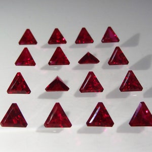 Può includere: Una collezione di 15 pietre preziose di rubino triangolari, ciascuna con un colore rosso intenso e un taglio sfaccettato. Le pietre preziose sono disposte in una griglia su una superficie bianca.