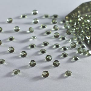 Peut inclure: Une collection de petites pierres précieuses rondes et facettées de couleur vert olive éparpillées sur une surface, avec une pelle en métal contenant un tas plus important de ces pierres étincelantes dans le coin supérieur droit.