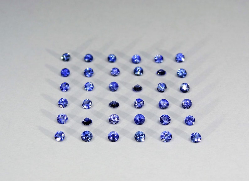 May include: A collection of thirty-six round, faceted blue gemstones arranged in a six-by-six grid pattern. Each individual stone displays a brilliant cut with multiple facets that reflect light, creating a uniform appearance across the set.