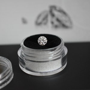 May include: A clear plastic container with a black velvet interior holds a round, clear diamond.