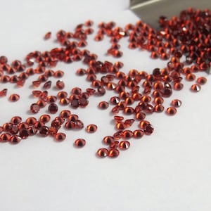 May include: A collection of small, round, faceted deep red gemstones scattered across a white surface. A metallic scoop is positioned at the top right, partially filled with more of these loose, brilliant-cut red stones, creating a vibrant contrast.