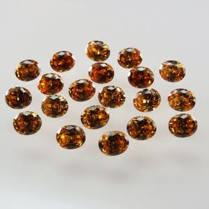 Pode incluir: Uma coleção de dezassete pedras preciosas soltas, com corte oval, numa cor âmbar-acastanhada profunda e rica. Cada pedra apresenta uma superfície facetada que reflete a luz, criando um aspeto cintilante nas gemas de tom castanho.
