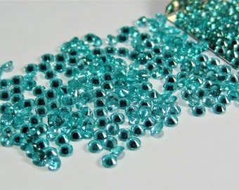 May include: A large collection of small, round, faceted turquoise-colored gemstones scattered across a surface, with a portion of the stones contained within a metallic scoop positioned at the top right corner of the frame.