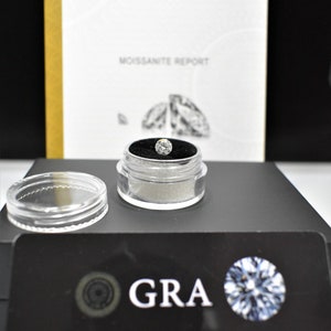 May include: A clear plastic container with a black lid holds a round, clear gemstone. The container is sitting on a black surface with a white label that reads "GRA Created by GRA Limited Lifetime Warranty and Certificate of Authentic REGISTER your GRA purchase Online www.gramosannite.com".