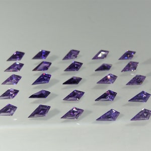 Peut inclure: Vingt-deux pierres précieuses facettées violettes disposées en rangées sur une surface blanche. Les pierres précieuses ont la forme de diamants allongés avec des extrémités pointues.