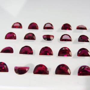 May include: A collection of 18 half-moon shaped ruby gemstones, arranged in rows on a white surface.