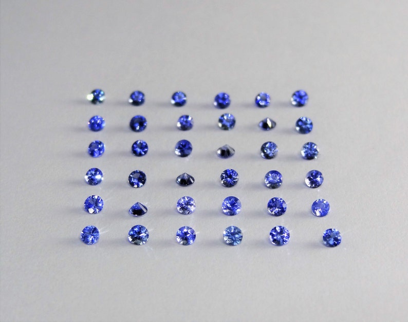 May include: A collection of thirty-six round, faceted blue gemstones arranged in a six-by-six grid. Each stone displays a brilliant cut with multiple facets that reflect light, showcasing a deep, vibrant blue color across the entire set.
