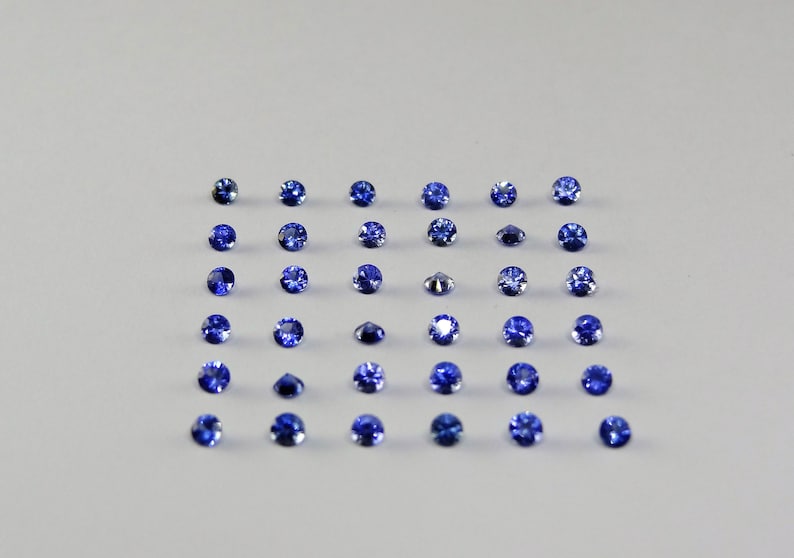 May include: A collection of thirty-six small, round, faceted blue gemstones arranged in a six-by-six grid. Each stone displays a deep, vibrant blue hue with reflective surfaces that catch the light, showcasing a uniform circular cut and polished finish.