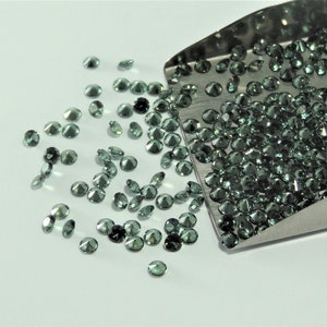 Può includere: Una collezione di piccole pietre preziose rotonde e sfaccettate color verde acqua scuro sparse accanto a una paletta in acciaio inossidabile riempita con una quantità maggiore delle stesse pietre circolari sciolte.