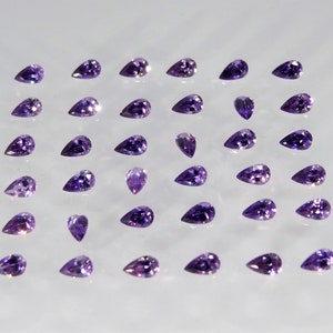 May include: A collection of 36 pear-shaped amethyst gemstones arranged in a grid pattern on a white surface.